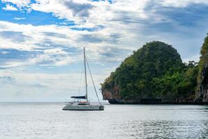 serene view of a sailboat anchored near a lush, green island with rocky cliffs under a bright, cloudy sky photo