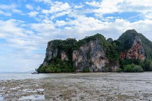 Towering cliffs covered in lush greenery, surrounded by a muddy beach after low tide with small boats in the distance, under a blue sky with scattered clouds photo