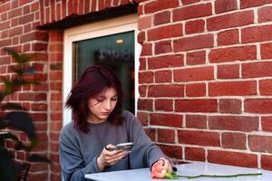 In a cozy cafe setting, a teenager with striking hair color rests her gaze on her mobile screen, a single rose on the table suggesting an introspective moment of solitude photo