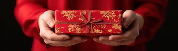 A person holding a beautifully wrapped red gift box adorned with gold patterns, symbolizing celebration and joy, against a dark background, creating a warm festive atmosphere. photo