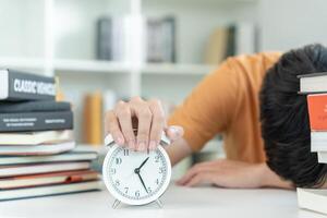 Asian student man turns off the alarm clock because he is tired from reading, male prepare for test and learning lessons in the library. stress, despair, haste, misunderstanding reading, discouraged. photo