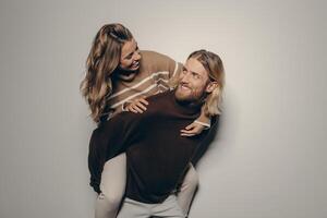 Joyful young elegant man piggybacking his beautiful girlfriend while standing on beige background photo