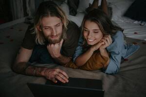 Happy young loving couple using laptop for net surfing while lying in bed at home together photo