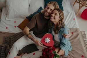 Top view of loving couple embracing and holding heart shape gift box while sitting on bed at home together photo