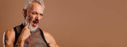 Playful mature gay man with rainbow colored eyebrows touching face with finger against brown background photo