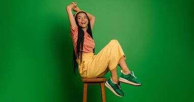 Beautiful young woman with dreadlocs looking happy while sitting on the chair against green background photo