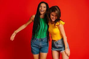 Two attractive young women in colorful wear embracing and smiling against red background photo