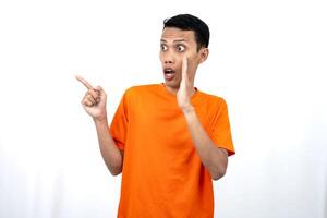 Portrait of a young Asian man wearing an orange t-shirt with his hand near his mouth in a whispering pose while pointing to the side of the copy space. Isolated on white background. photo