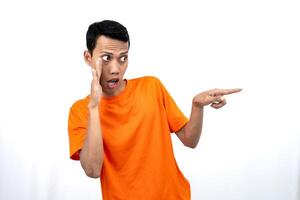 Portrait of a young Asian man wearing an orange t-shirt with his hand near his mouth in a whispering pose while pointing to the side of the copy space. Isolated on white background. photo