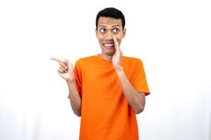 Portrait of a young Asian man wearing an orange t-shirt with his hand near his mouth in a whispering pose while pointing to the side of the copy space. Isolated on white background. photo