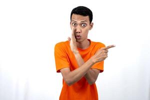 Portrait of a young Asian man wearing an orange t-shirt with his hand near his mouth in a whispering pose while pointing to the side of the copy space. Isolated on white background. photo