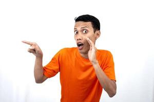 Portrait of a young Asian man wearing an orange t-shirt with his hand near his mouth in a whispering pose while pointing to the side of the copy space. Isolated on white background. photo