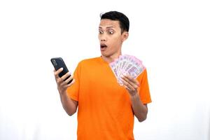 Portrait of a young Asian man wearing an orange t-shirt with a happy, surprised, pleased expression while carrying a smartphone and hundreds of thousands of rupiah. isolated on a white background. photo