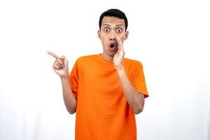 Portrait of a young Asian man wearing an orange t-shirt with his hand near his mouth in a whispering pose while pointing to the side of the copy space. Isolated on white background. photo
