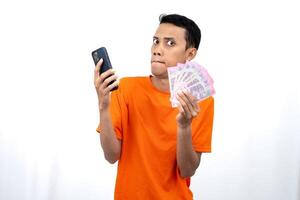 Portrait of a young Asian man wearing an orange t-shirt with a happy, surprised, pleased expression while carrying a smartphone and hundreds of thousands of rupiah. isolated on a white background. photo