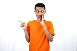 Portrait of a young Asian man wearing an orange t-shirt with his hand near his mouth in a whispering pose while pointing to the side of the copy space. Isolated on white background. photo