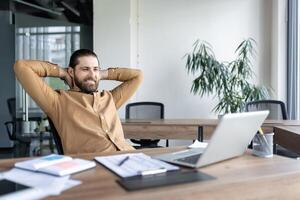 Confident businessman seated at desk with hands behind head, smiling while taking break. Modern office environment with natural light, laptop, and documents reflecting comfortable workplace. photo