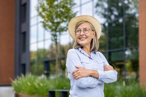 Senior woman wearing glasses and straw hat stands confidently with arms crossed outside modern office building. Scene captures happiness, positivity, and elegance photo