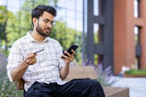Man sitting on bench outdoors holding credit card and phone with concerned expression, checking for possible fraud or online transaction issues photo