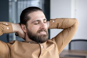 Man with beard enjoying moment of relaxation in office setting. Hands behind head, expressing comfort and satisfaction. Casual shirt, confident expression symbolizes stress-free atmosphere photo