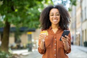 Smiling woman using smartphone for online shopping with credit card outdoors. Digital payment concept shown through confident, happy expression and modern technology use in urban setting. photo