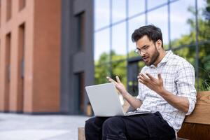 Young man shows frustration while working on laptop outdoors. Urban setting with modern office buildings in background. Difficulties with technology, work stress, and problem solving portrayed photo