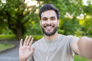 Joyful young man taking selfie outdoors, smiling and waving while enjoying music on earphones. Captures happiness and connection in natural setting with green trees, portraying positive communication photo