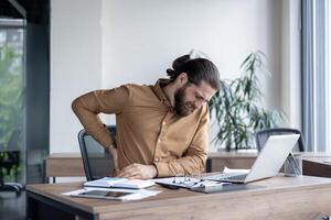 Man in office setting suffers from back pain while working on laptop. Displays discomfort at work desk, surrounded by documents, glasses, and modern office furniture. photo