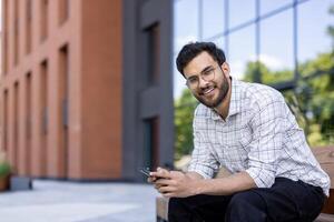 Man wearing glasses sitting on bench outside modern building holding smartphone. Smiling relaxed individual enjoying outdoor environment with blurred urban architecture background. C photo