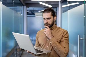 A thoughtful male programmer holding a laptop and analyzing data in a contemporary office setting. The image captures a moment of concentration and decision-making. photo