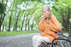 Senior woman sitting on park bench struggles with breathing. Image captures concern and health issues in tranquil natural environment. Depicts aging, respiratory problems, need for medical attention. photo