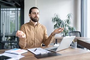 Relaxed professional man meditating at work desk to find balance and reduce stress. Seated in modern office environment, with laptop and documents, embracing mindfulness during work hours. photo