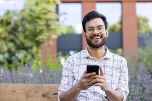 Close-up portrait of a young Indian man wearing glasses and a shirt sitting on a bench outside a building, holding a mobile phone and smiling at the camera. photo