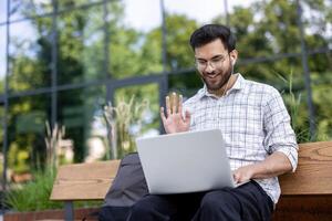 Smiling man using laptop outdoors holding call with headphones wearing casual attire. Background of modern building , emphasizing connection, technology, and remote communication photo