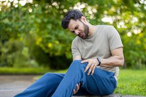 Man holds sore knee with expression of discomfort while sitting on grass during outdoor exercise. Scene captures moment of physical challenge, highlighting need for rest and recovery. photo