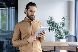Businessman stands in modern office using digital tablet, contemplating business strategy surrounded by natural light and greenery. Image conveys focus, technology, and professional atmosphere photo