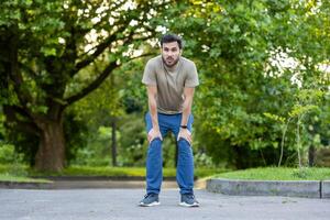 Young sportsman resting after an intense exercise session in park. Dressed in casual sportswear, he leans forward catching breath, surrounded by serene green environment. photo