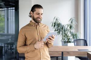 Young man with beard holding tablet in office setting. Smiling, engaged with digital device, reflecting modern business communication style. Office workspace hints collaboration and technology-driven photo