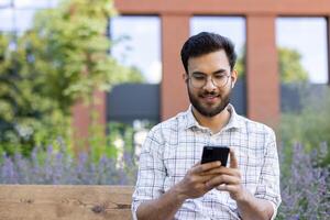 Smiling man sitting on bench using smartphone in urban outdoor setting with greenery. Enjoying technology and communication. photo