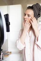 Blogger recording skincare routine. Woman applying eye patches in bathroom in front of phone photo