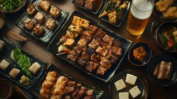 A high-angle shot of a Japanese izakaya table filled with small dishes of grilled meats, tofu, pickles, and a glass of cold beer in the corner of the frame photo