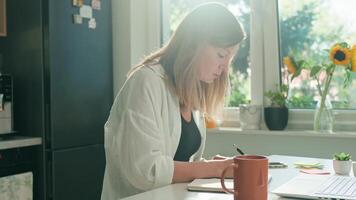 Woman is working remotely at desk in home office, taking notes in notebook. Back view of female freelancer using laptop in the morning video