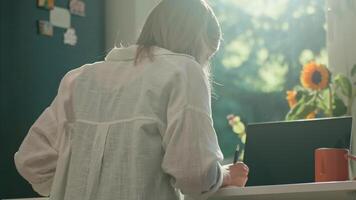 Woman is working remotely at desk in home office, taking notes in notebook. Back view of female freelancer using laptop in the morning video