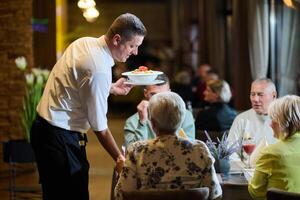 A courteous waiter provides impeccable service to an elderly family and their friends, creating a convivial atmosphere as they gather for a shared lunch, indulging in the diverse and delectable photo