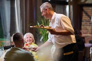 A courteous waiter provides impeccable service to an elderly family and their friends, creating a convivial atmosphere as they gather for a shared lunch, indulging in the diverse and delectable photo