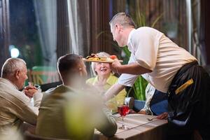 A courteous waiter provides impeccable service to an elderly family and their friends, creating a convivial atmosphere as they gather for a shared lunch, indulging in the diverse and delectable photo