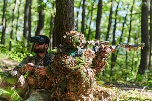A skilled sniper and a soldier operating a drone with VR goggles strategize and observe the military action while concealed in the forest photo