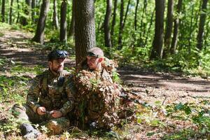 A skilled sniper and a soldier operating a drone with VR goggles strategize and observe the military action while concealed in the forest photo