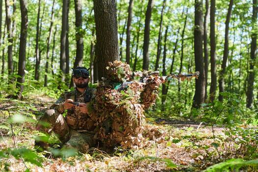A skilled sniper and a soldier operating a drone with VR goggles strategize and observe the military action while concealed in the forest photo