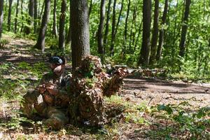 A skilled sniper and a soldier operating a drone with VR goggles strategize and observe the military action while concealed in the forest photo
